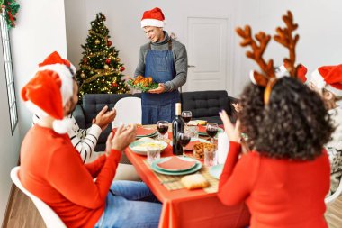 Bir grup insan el çırpıyor ve masada oturuyor. Ayakta duran ve elinde kavrulmuş hindi tutan adam Noel 'i evde kutluyor..