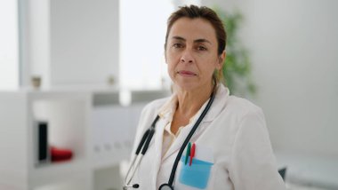 Orta yaşlı İspanyol kadın doktor üniforması giyiyor klinikte rahat bir ifadeyle dikiliyor.