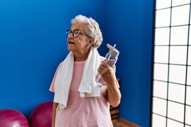 Spor kıyafetleri giymiş, spor salonunda elinde su şişesi tutan yaşlı, kır saçlı bir kadın.
