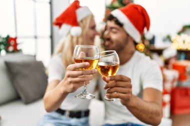 Mutlu bir şekilde gülümseyen genç çift Noel şapkası takıyor evde şampanyayla kadeh kaldırıyor.