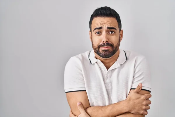 Young hispanic man with beard wearing casual clothes over white background shaking and freezing for winter cold with sad and shock expression on face