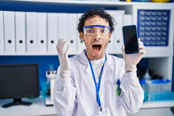 Hispanic man working at scientist laboratory showing smartphone screen celebrating victory with happy smile and winner expression with raised hands