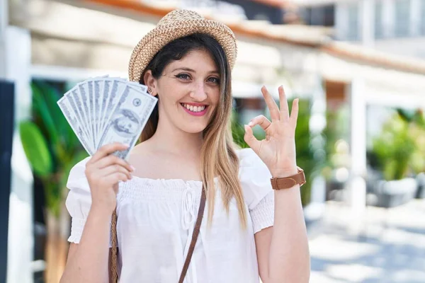 Młoda Brunetka Trzyma Banknoty Dolarów Robi Znak Palcami Uśmiechając Przyjazny — Zdjęcie stockowe