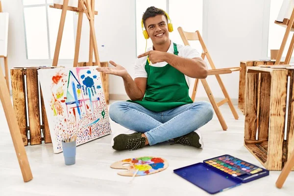 Young hispanic man at art studio amazed and smiling to the camera while presenting with hand and pointing with finger.