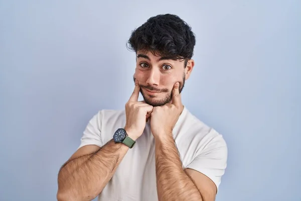 Hispanic man with beard standing over white background smiling with open mouth, fingers pointing and forcing cheerful smile