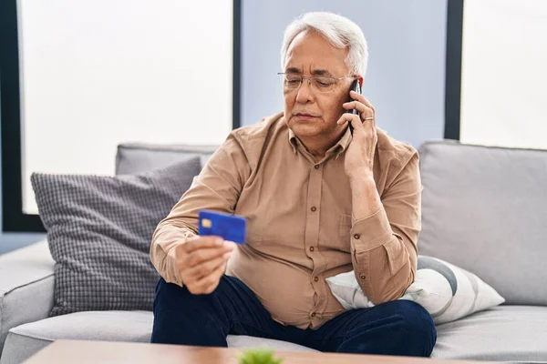 Senior man talking on the smartphone holding credit card sitting on sofa at home