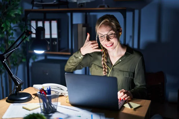 Young blonde woman working at the office at night smiling doing phone gesture with hand and fingers like talking on the telephone. communicating concepts.