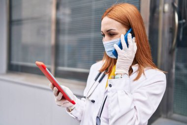 Tıbbi maske takan, dokunmatik ped kullanan beyaz kadın doktor hastanede akıllı telefondan konuşuyor.