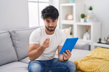 Hispanic man with beard using touchpad sitting on the sofa doing money gesture with hands, asking for salary payment, millionaire business 