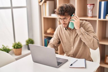 Genç iş adamı ofisteki bilgisayarı kullanarak akıllı telefondan konuşuyor.