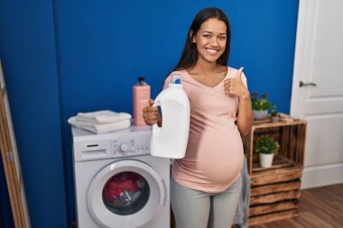 Çamaşır deterjanı şişesini tutan hamile bir kadın mutlu ve pozitif gülümsüyor, başparmağı mükemmel ve onay işareti yapıyor. 