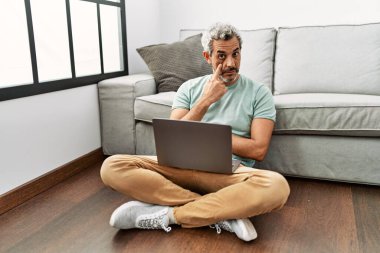 Orta yaşlı İspanyol adam oturma odasında dizüstü bilgisayar kullanıyor. Gözünü dikip jestini izliyor. Şüpheli bir ifade. 