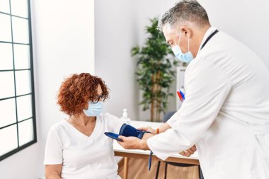 Klinikte gerilim ölçer kullanarak doktor üniforması ve tıbbi maske takan orta yaşlı bir adam ve kadın.