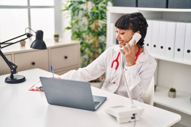 Doktor üniforması giyen genç bir kadın klinikte telefonla konuşuyor.
