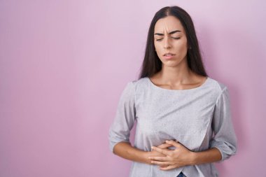 Genç esmer kadın, elleri karnında pembe arka planda duruyor çünkü hazımsızlık, acı verici bir hastalık kendini iyi hissetmiyor. Ağrı kavramı. 