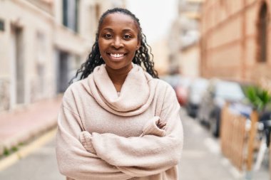 Afro-Amerikalı kadın gülümsüyor. Kollarını kavuşturup caddede duruyor.