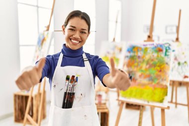 Sanat stüdyosundaki genç esmer kadın elleriyle olumlu bir jest yapmayı onaylıyor, gülümsüyor ve başarı için mutlu. kazanan jest. 