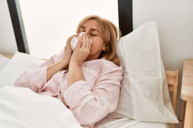 Orta yaşlı sarışın kadın mutlu bir şekilde gülümsüyor. Evdeki yatakta yatıyor..