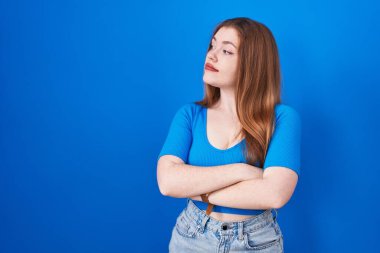 Mavi arka planda duran kızıl saçlı kadın kollarını kavuşturmuş, ikna olmuş ve kendinden emin bir şekilde yan tarafa bakıyor. 