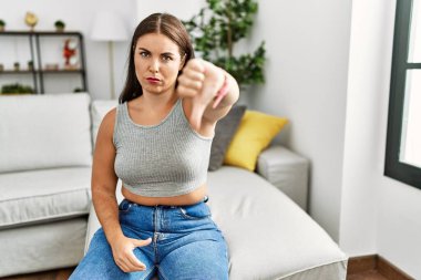 Evdeki kanepede oturan genç esmer kadın mutsuz ve kızgın görünüyor. Reddedilmiş ve olumsuz hareketler yapıyor. kötü ifade. 