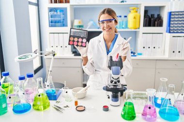Young blonde woman working at scientist laboratory with make up smiling happy pointing with hand and finger to the side 