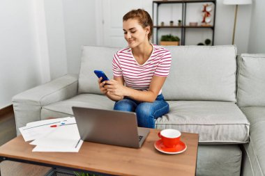 Akıllı telefon ve dizüstü bilgisayar kullanan genç bir kadın evdeki kanepede oturuyor.