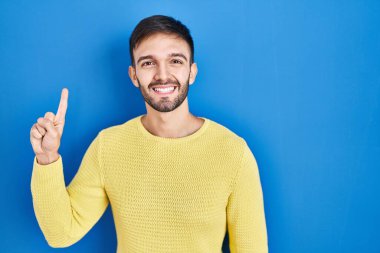 Mavi arka planda duran İspanyol adam bir numarayı gösterip işaret ederken kendinden emin ve mutlu gülümsüyor.. 