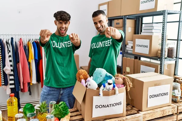Young gay couple wearing volunteer t shirt at donations stand pointing to you and the camera with fingers, smiling positive and cheerful