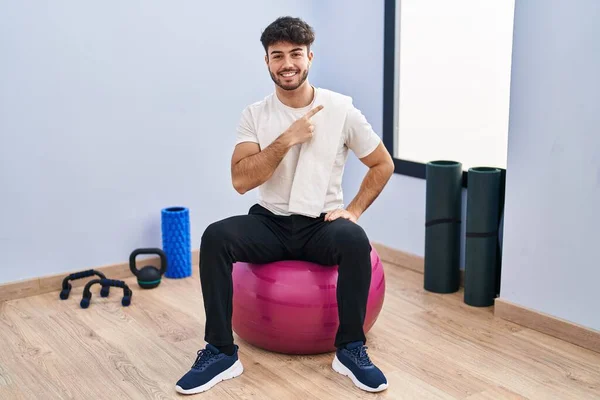 Hispanic man with beard sitting on pilate balls at yoga room cheerful with a smile of face pointing with hand and finger up to the side with happy and natural expression on face