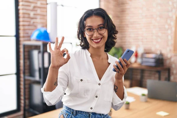 スマートフォンを使ってオフィスで働く若いヒスパニック系の女性は 指でOkサインを行います 笑顔優しいジェスチャー優れたシンボル — ストック写真