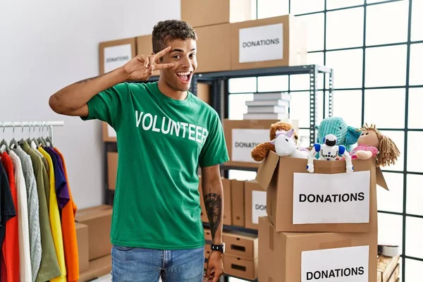 Young handsome hispanic man wearing volunteer t shirt at donations stand doing peace symbol with fingers over face, smiling cheerful showing victory