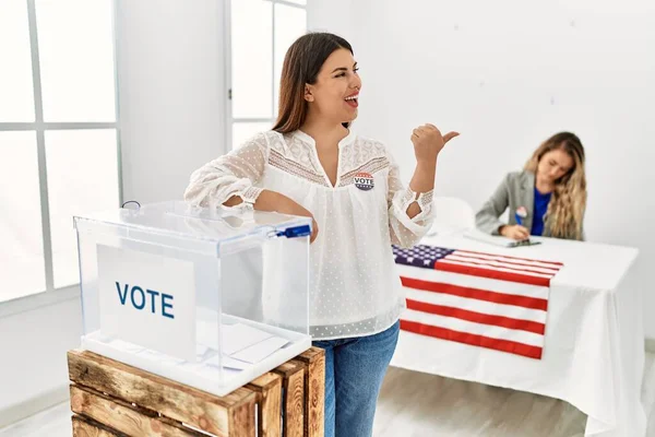 Young brunette woman voting putting envelop in ballot box pointing thumb up to the side smiling happy with open mouth