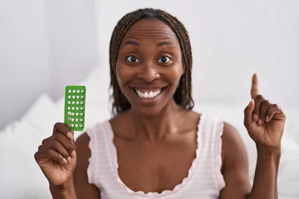 African american woman holding birth control pills surprised with an idea or question pointing finger with happy face, number one