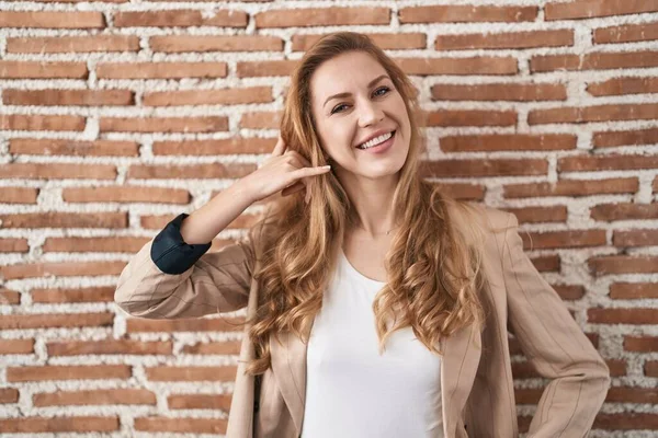 Beautiful blonde woman standing over bricks wall smiling doing phone gesture with hand and fingers like talking on the telephone. communicating concepts.