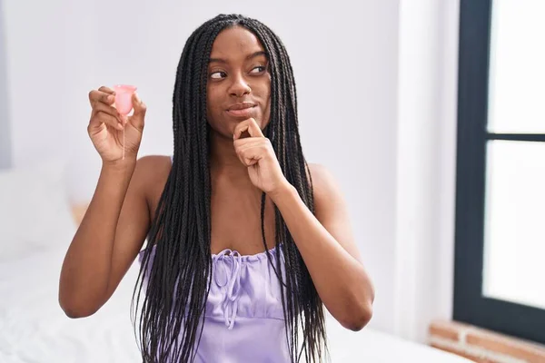 Young african american with braids holding menstrual cup serious face thinking about question with hand on chin, thoughtful about confusing idea