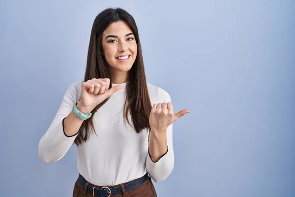 Young brunette woman standing over blue background pointing to the back behind with hand and thumbs up, smiling confident