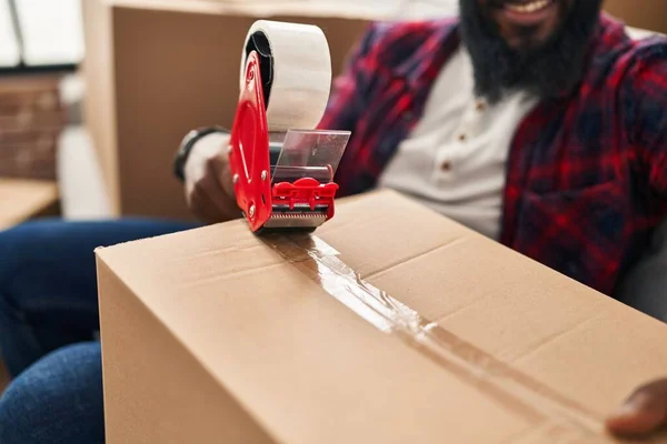 Young african american man packing cardboard box sitting on sofa at new home