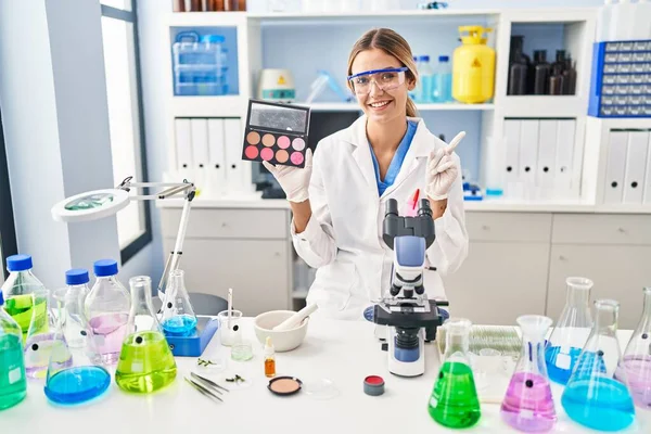 Young blonde woman working at scientist laboratory with make up smiling happy pointing with hand and finger to the side 