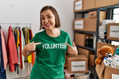 Beautiful middle age hispanic woman wearing volunteer t shirt at donations stand smiling happy pointing with hand and finger 