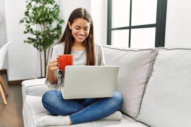 Young latin woman using laptop and drinking coffee sitting on sofa at home