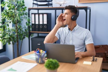 Young hispanic man working at the office wearing headphones shouting and screaming loud to side with hand on mouth. communication concept. 
