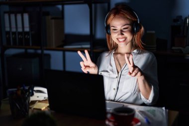 Young caucasian woman working at the office at night smiling looking to the camera showing fingers doing victory sign. number two. 