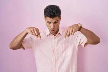 Young hispanic man standing over pink background pointing down with fingers showing advertisement, surprised face and open mouth 