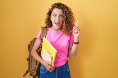 Sırt çantası takan ve elinde başarılı bir fikirle işaret eden kitaplar tutan beyaz bir kadın. Heyecanlı ve mutlu. Bir numara.. 