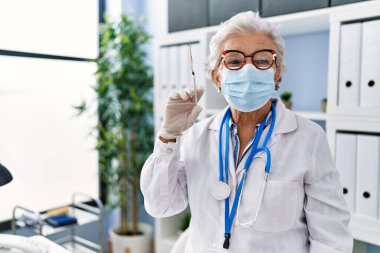 Gri saçlı, doktor üniforması giyen ve elinde şırınga tutan yaşlı bir kadın. Pozitif ve mutlu görünüyor. Kendine güvenen bir gülümsemeyle dişlerini gösteriyor. 