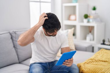 Hispanic man with beard using touchpad sitting on the sofa confuse and wondering about question. uncertain with doubt, thinking with hand on head. pensive concept. 