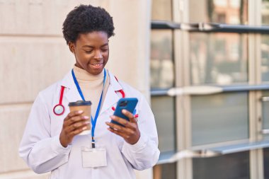 Afro-Amerikan kadın doktor üniforması giyiyor akıllı telefon kullanıyor sokakta kahve içiyor.