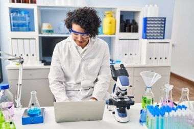 Laboratuvarda dizüstü bilgisayar ve mikroskop kullanan bilim adamı üniforması giyen genç bir İspanyol.