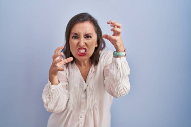 Middle age hispanic woman standing over blue background shouting frustrated with rage, hands trying to strangle, yelling mad 