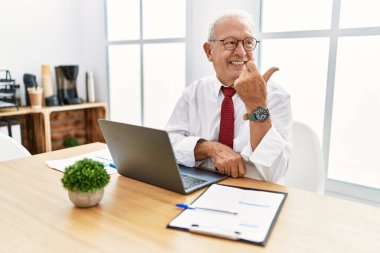 Senior man working at the office using computer laptop smiling with happy face looking and pointing to the side with thumb up. 